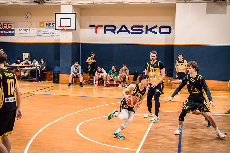 Hráči basketbalu během zápasu v tělocvičně s logem TRASKO na stěně.